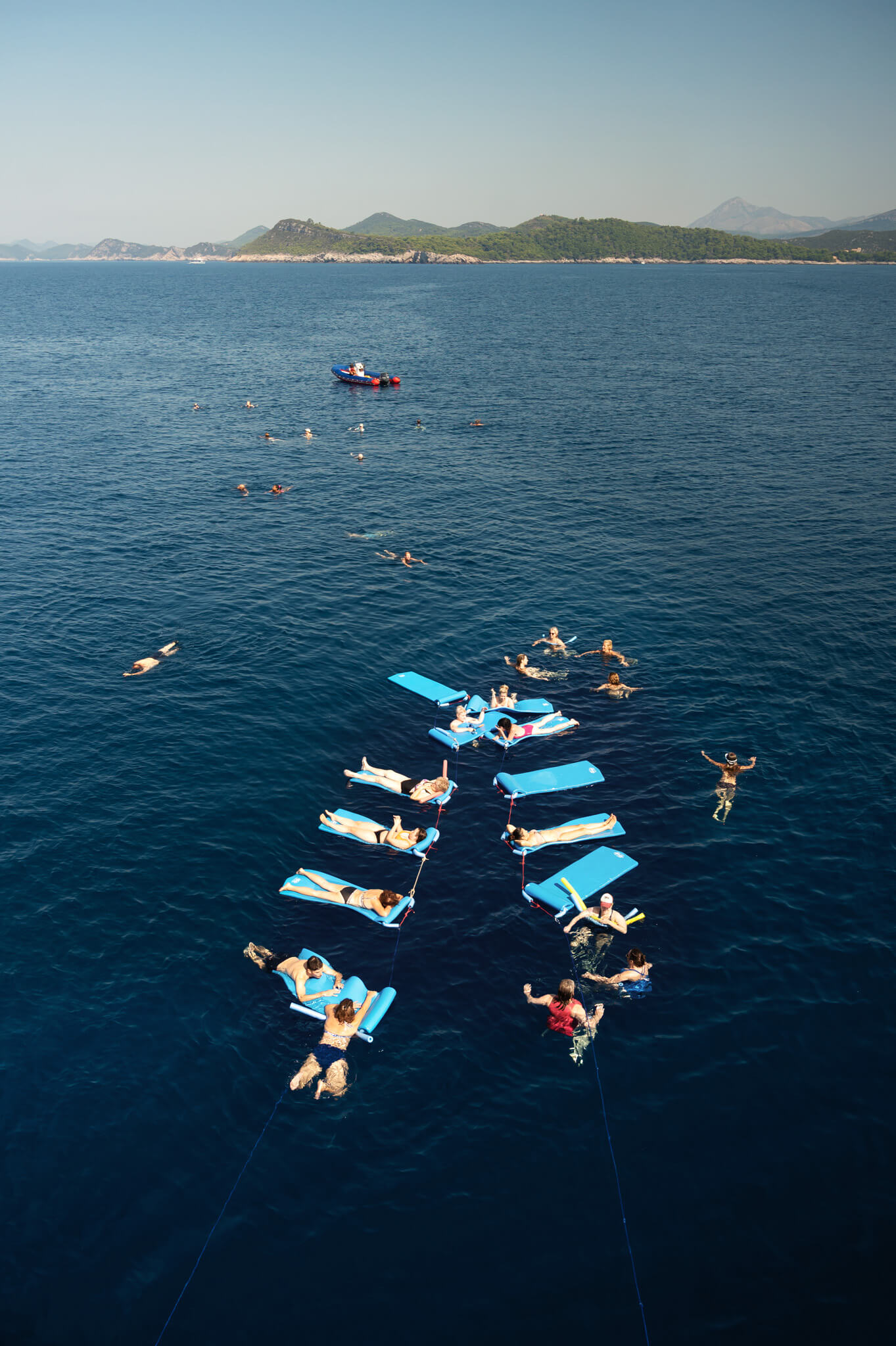 Star Clippers Royal Clipper Ship 18 • Our Little Lifestyle Aerial view of people lounging on bright blue inflatable mats tethered together in the sea near a rocky coastline with green hills in the distance.
