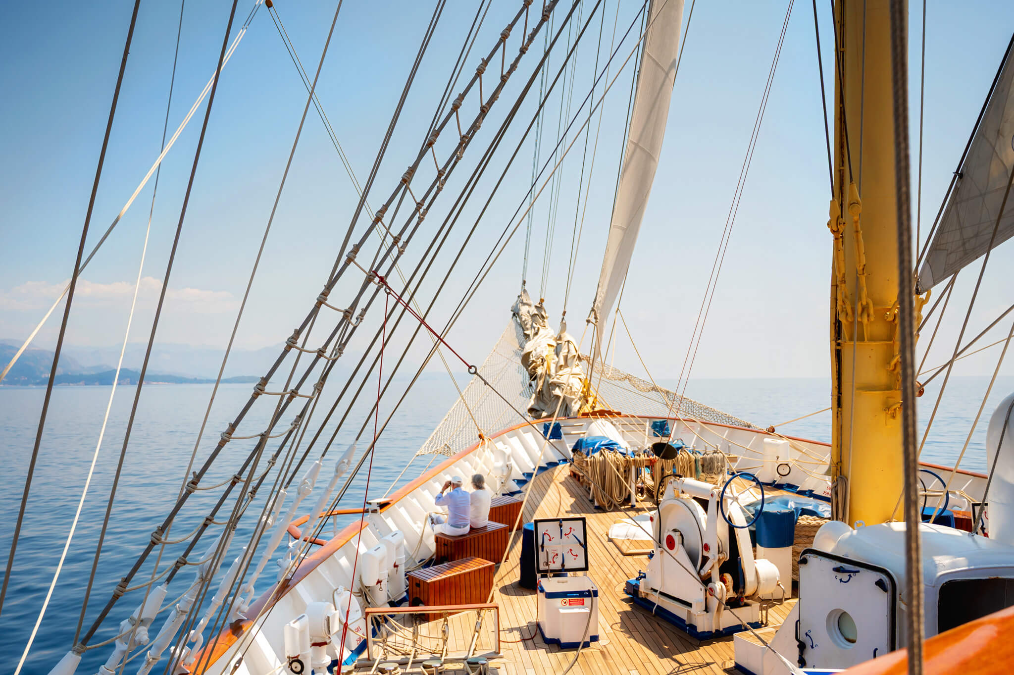 Sailing vessel deck with numerous rigging lines crossing the frame, a yellow mast on the right, and a calm blue sea and sky in the background.