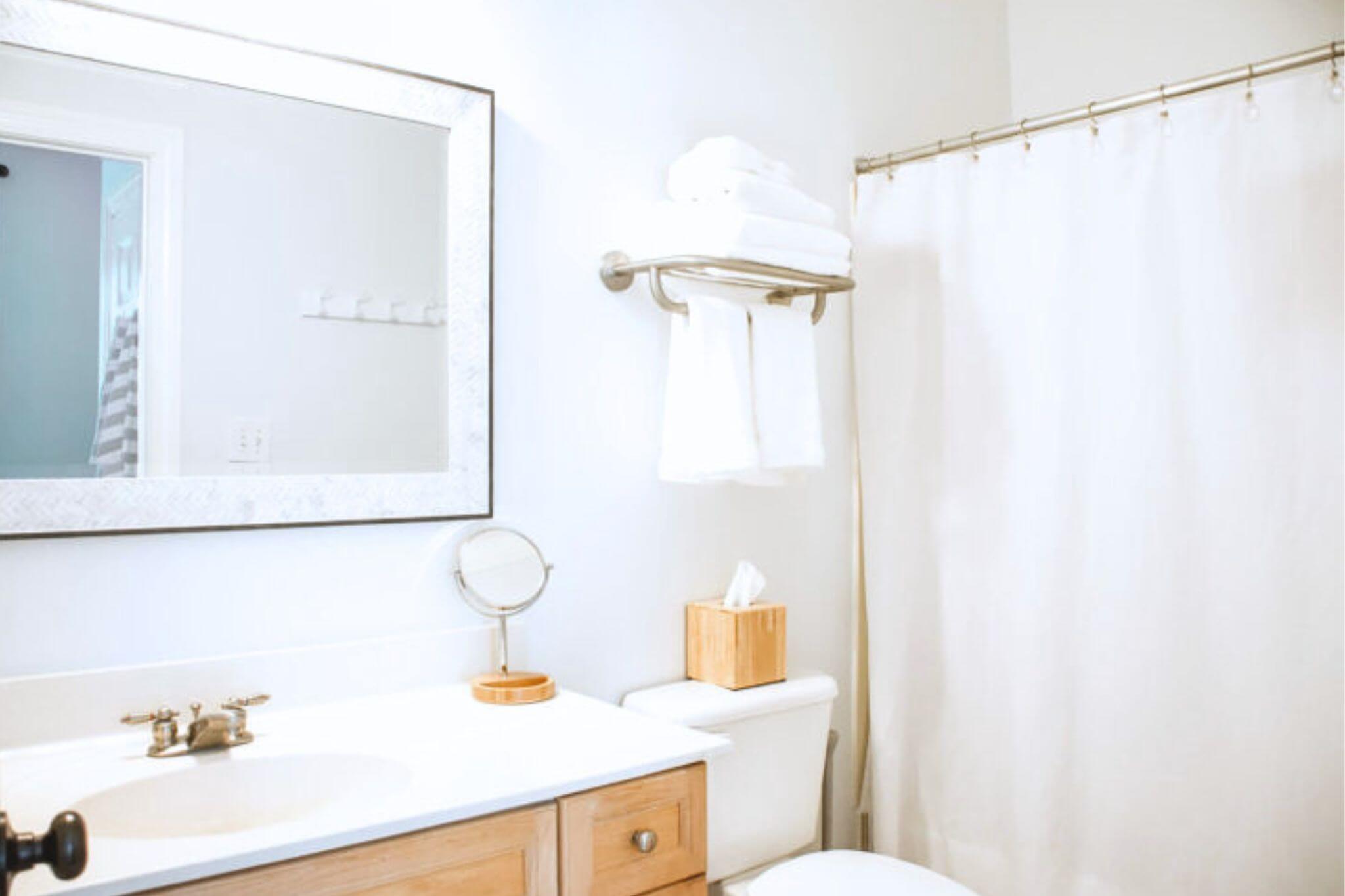 Bathroom with a white sink and wooden vanity, large mirror, and a stack of white towels on a wall rack by the shower.
