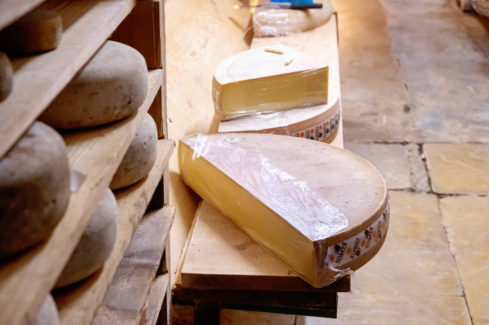 Wheel of Comte cheese wrapped in plastic on wooden shelves
