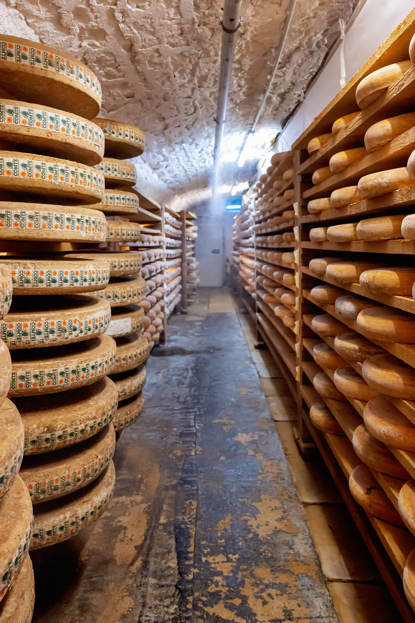 Narrow cellar aisle with large rounds of comte cheese stacked on wooden racks along both walls