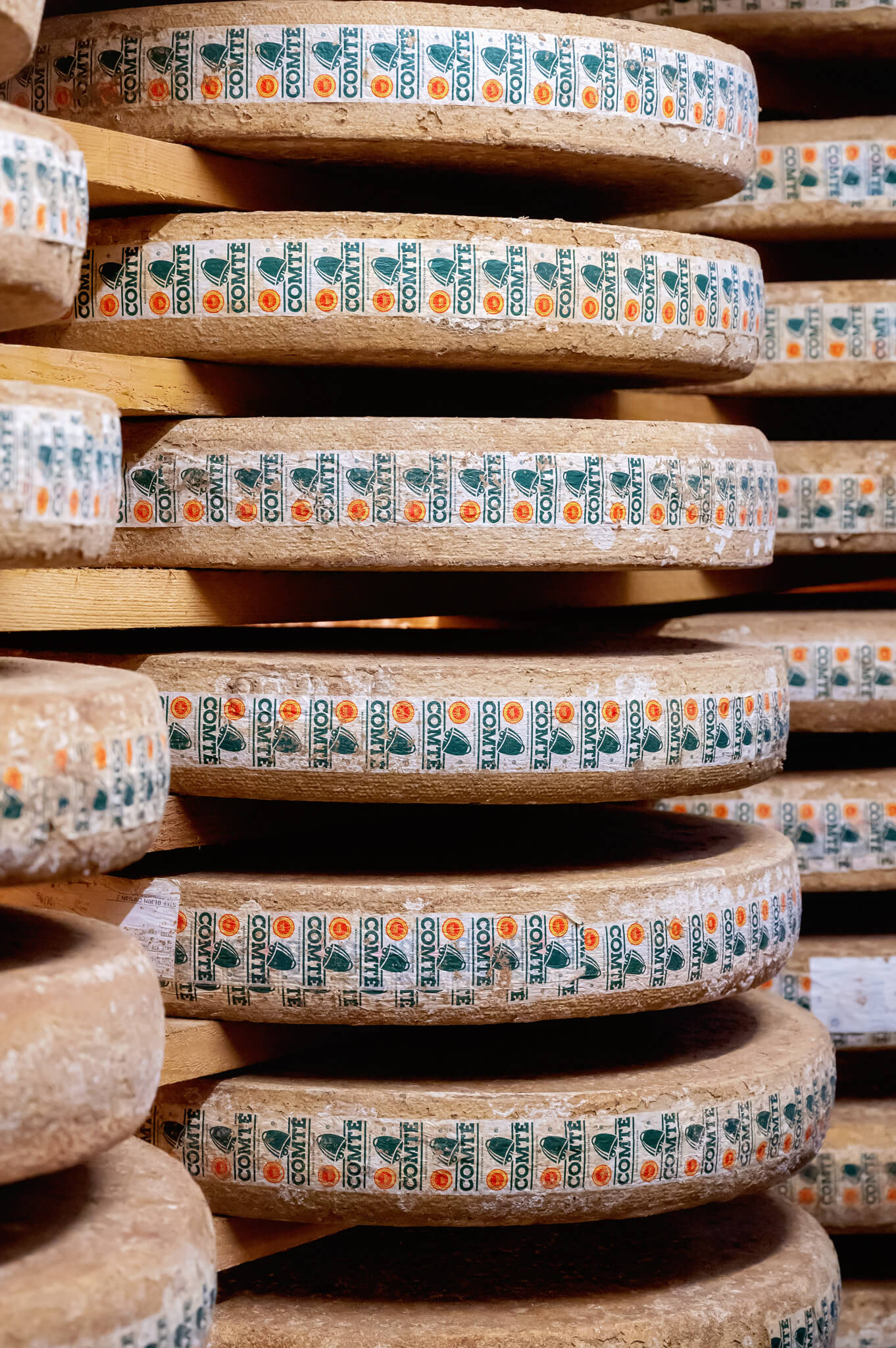 Stack of round comte cheese wheels with rough brown rind, wrapped in repeating COMTE labels, stacked on wooden shelves.