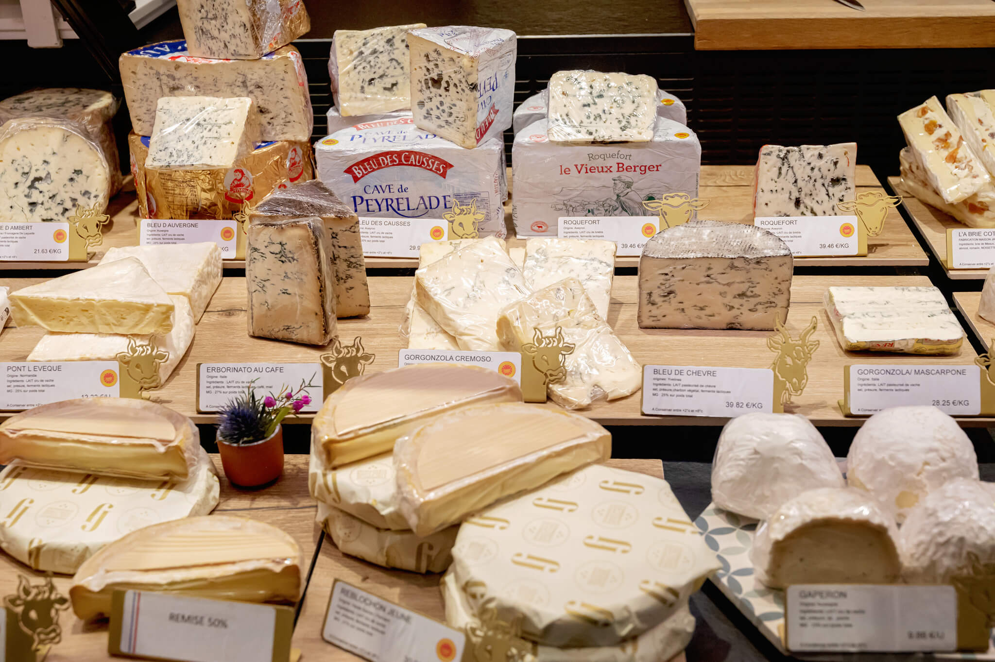 Assorted blue cheeses and wheel cheeses on a market-style display with labeled price tags and decorative cow icons