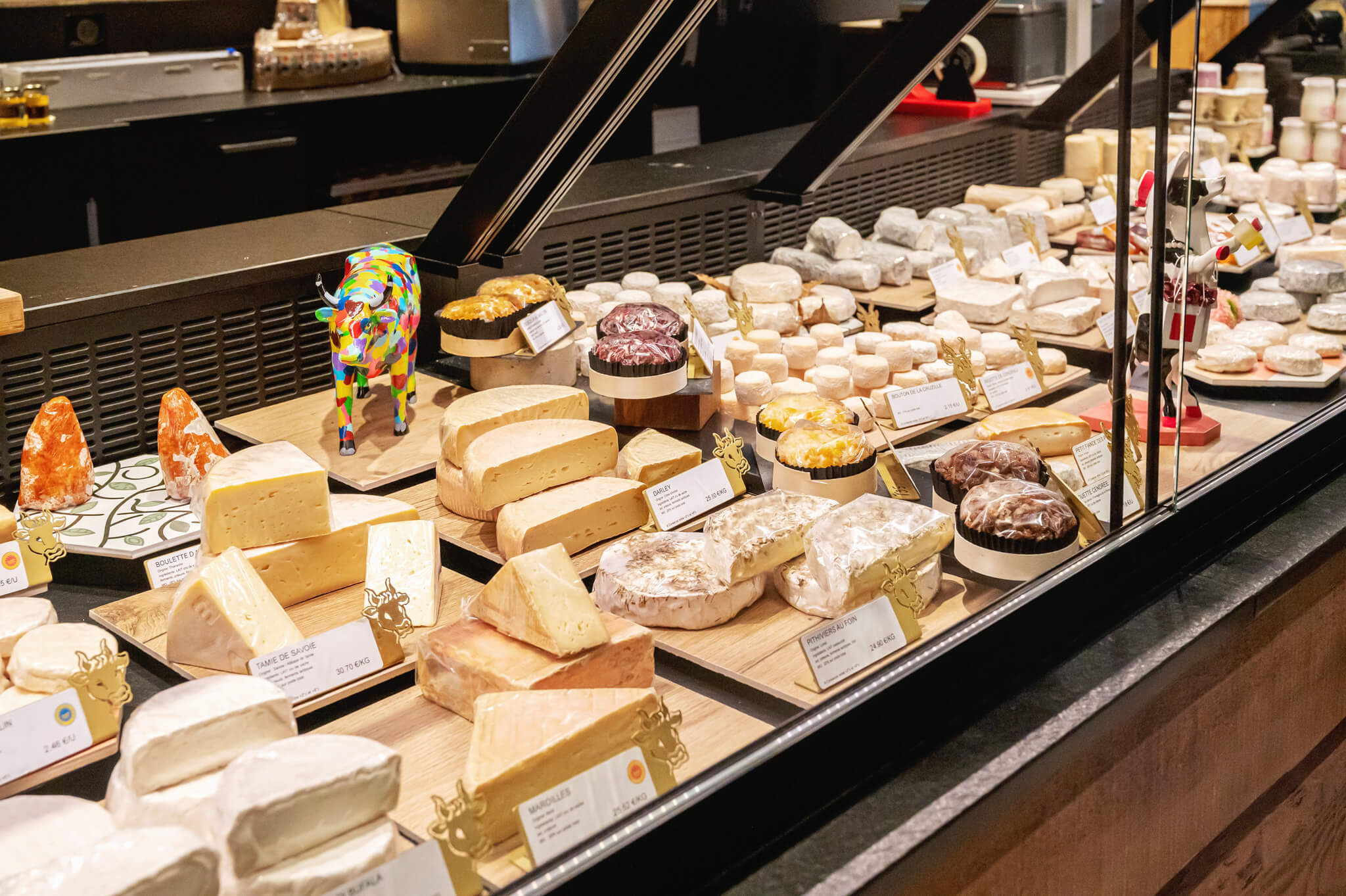 Assorted cheeses on display in a shop window, with wheels, wedges, and small price tags; a colorful decorative cow statue stands among the cheeses.