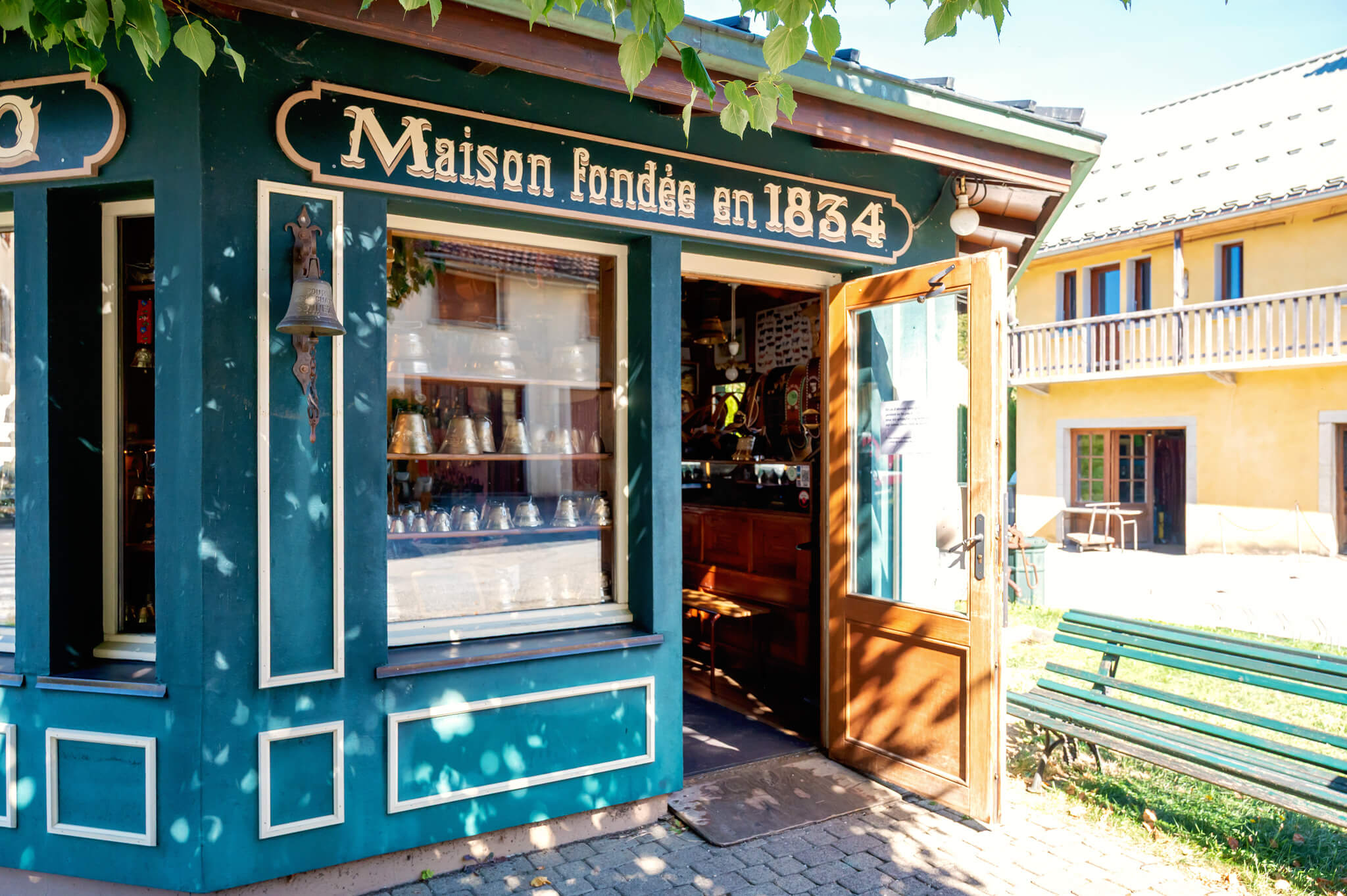 Teal storefront with white trim, an open wooden door, and a window display of bells; sign reads'Maison fondée en 1834' on the facade.