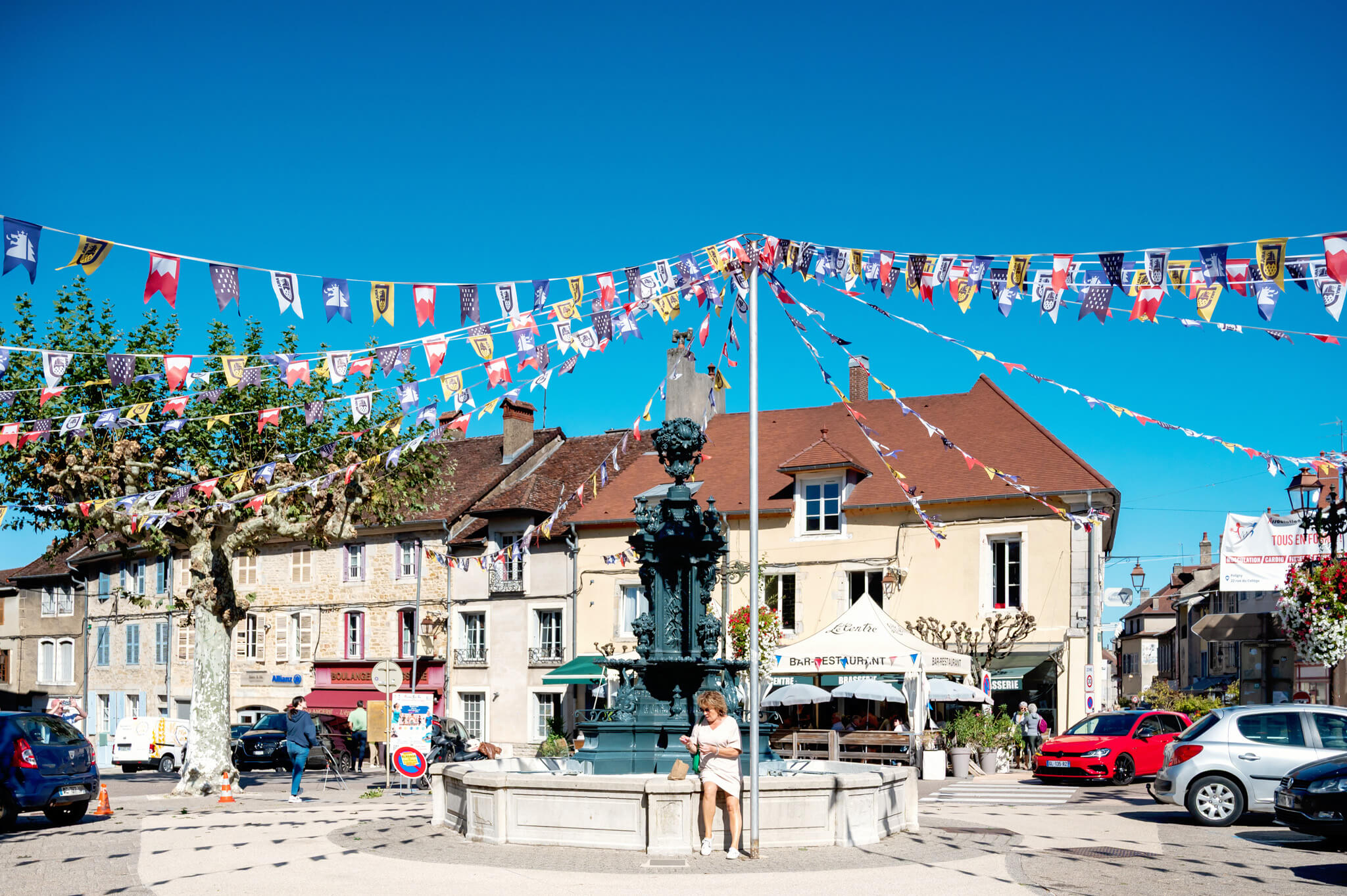 Public square with colorful triangular bunting overhead, ornate fountain in the center, and historic buildings beneath a clear blue sky.