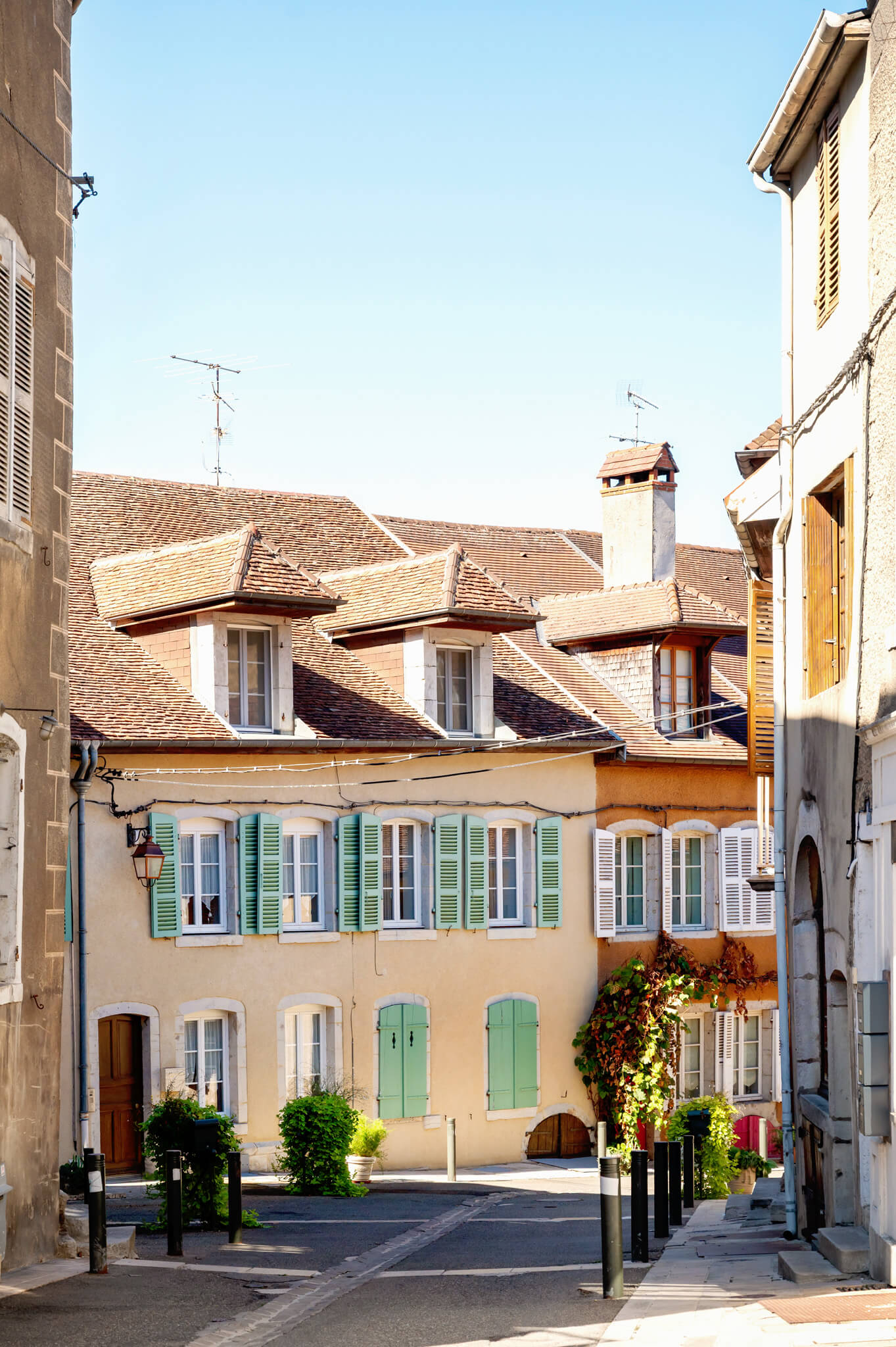 Colorful French alley with beige townhouses, green shutters, and clay-tiled roofs under a clear blue sky, leading through a small street lined with bollards and potted plants.
