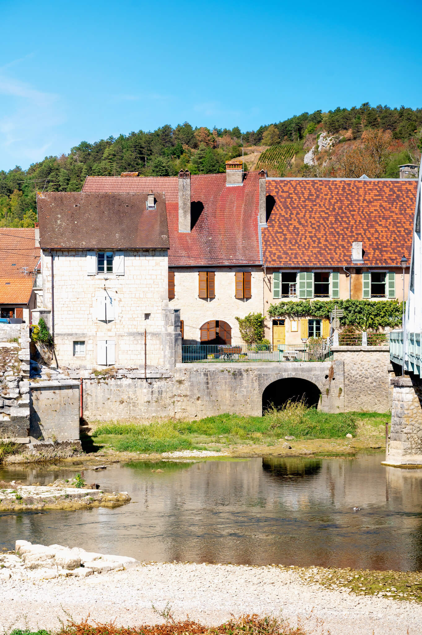 Row of stone houses with red tile roofs along a calm river, hillside with trees in the background.