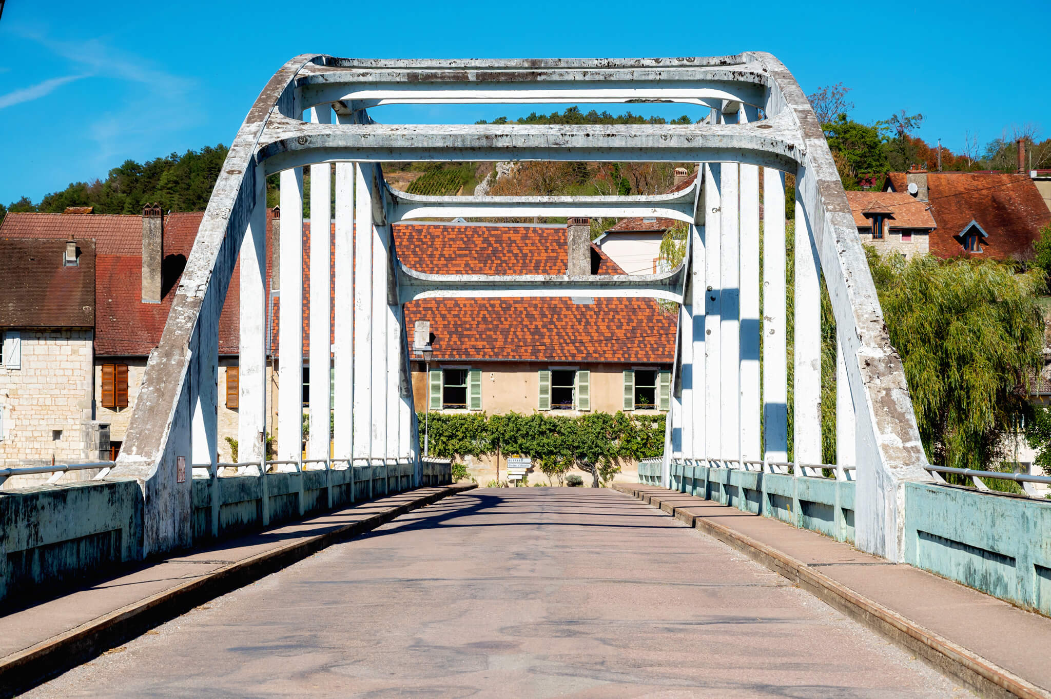 White weathered iron arch bridge seen from its center, leading to red-tiled buildings under a bright blue sky.
