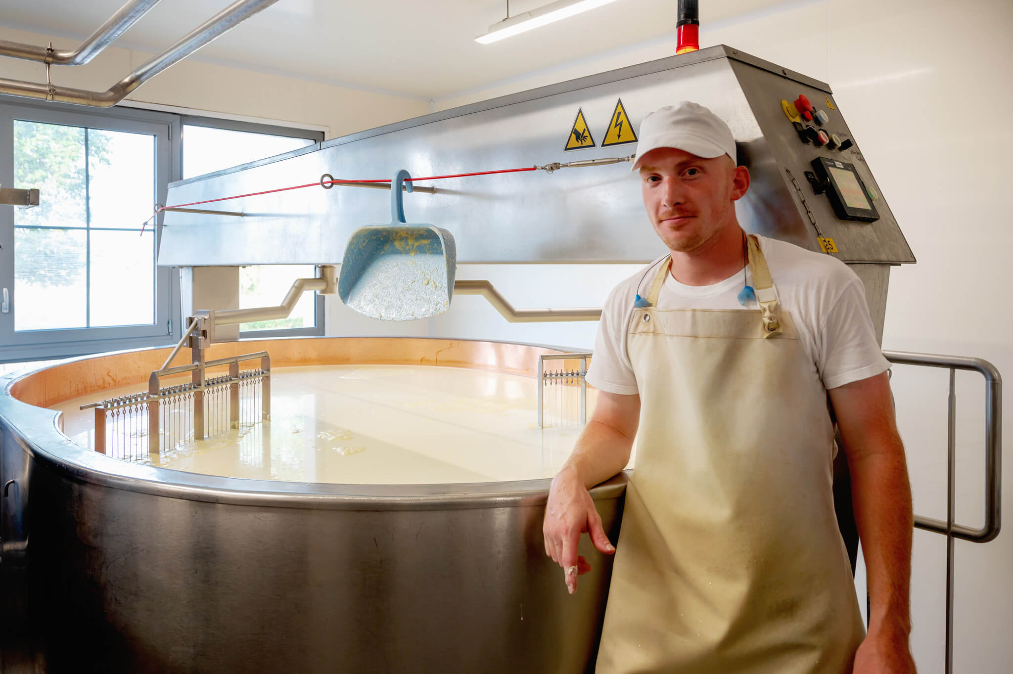 Male dairy plant worker in a white cap and apron beside a large stainless steel mixing vat in a dairy processing room, with a ladle hanging above it.