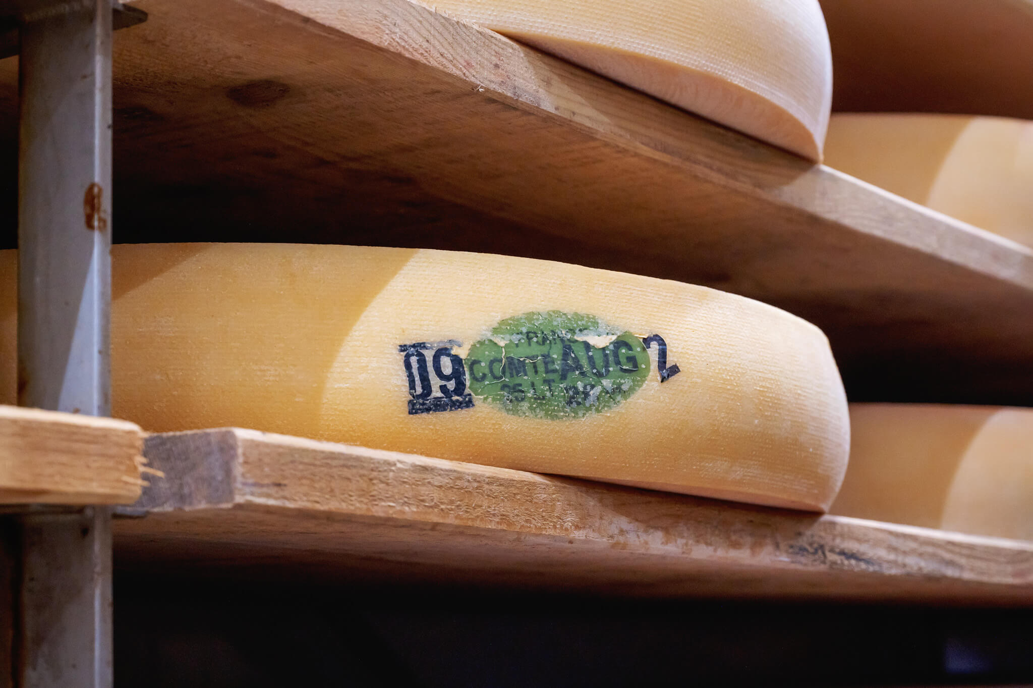 Close-up of stacked wooden boards on a shelf, with a worn green oval comte cheese stamp partially visible.