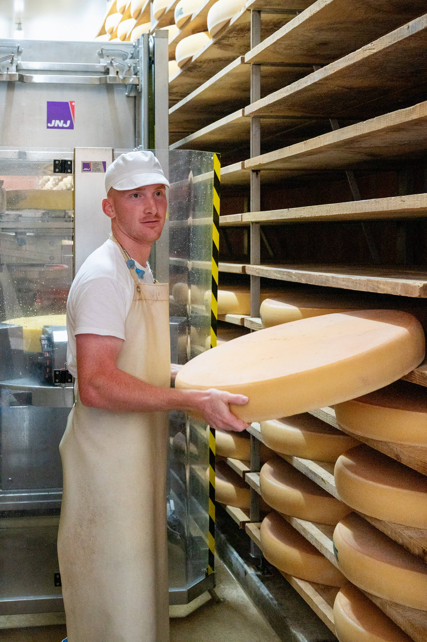Assistant cheese maker in a white hat and apron holding a large cheese wheel in a cheese-aging room with wooden shelves full of rounds nearby.