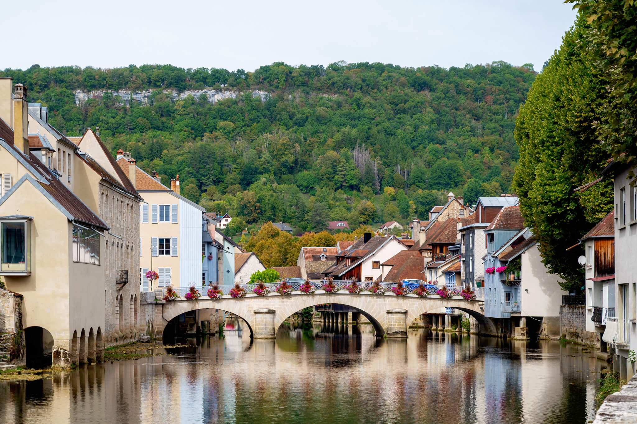 Ornans, France Town by a calm river with a stone-arched bridge and colorful houses, backed by a forested hill.