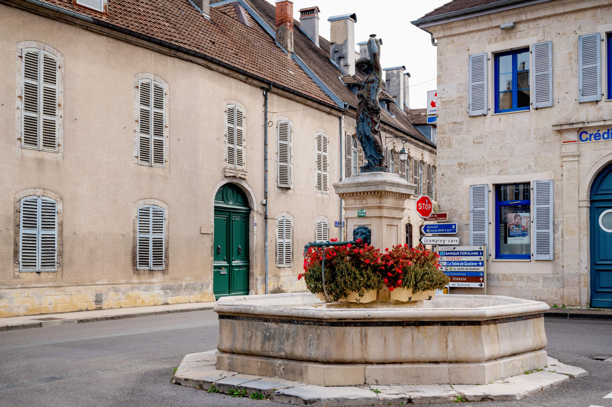 Ornans, France Circular stone fountain with a statue in a French town square, surrounded by beige buildings with shuttered windows and blue-framed doors and windows; red flowers in a planter nearby