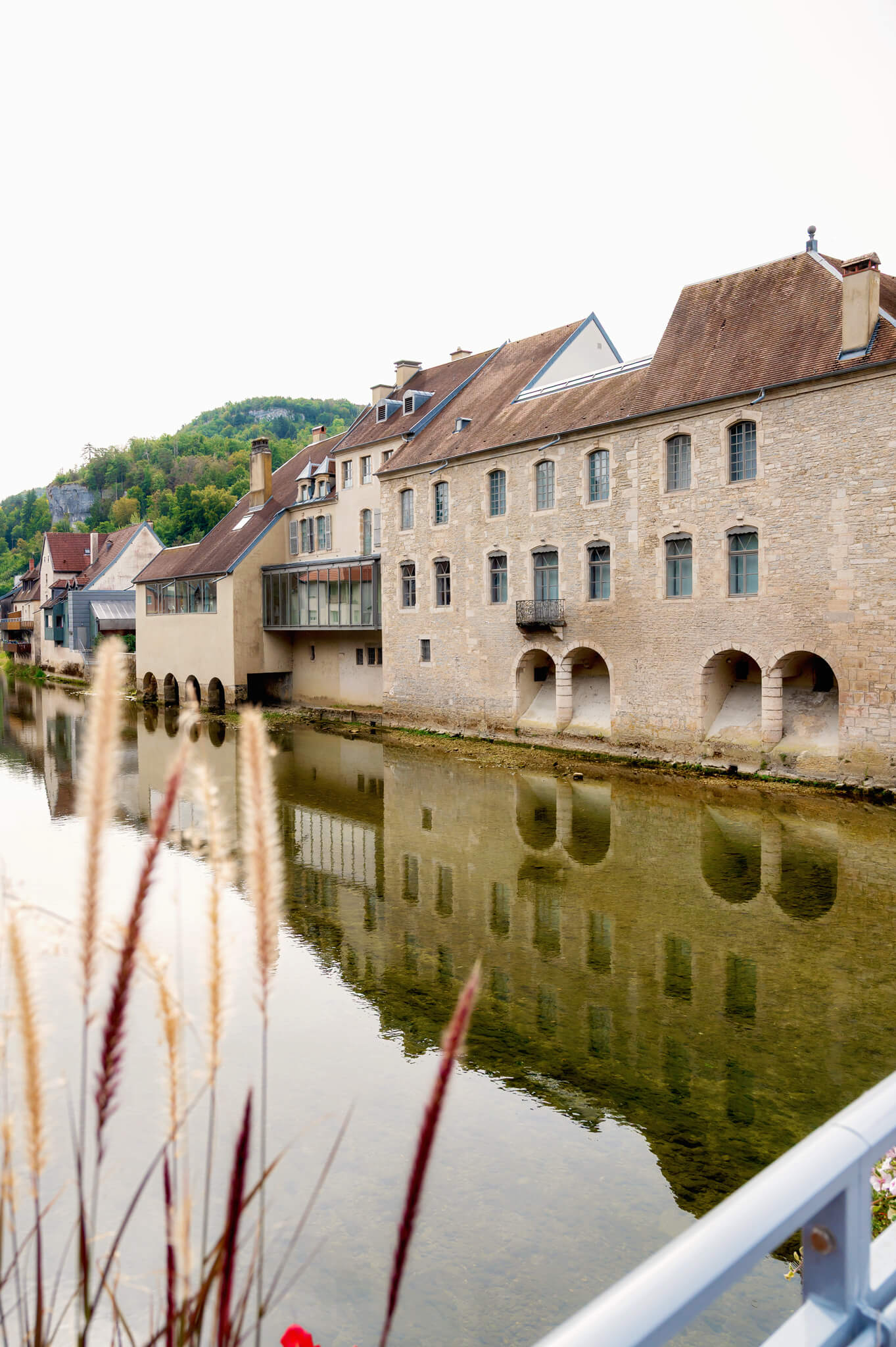 Ornans, France Stone buildings along a river with arched openings on the ground floor and their reflections in the water, hillside in the background.