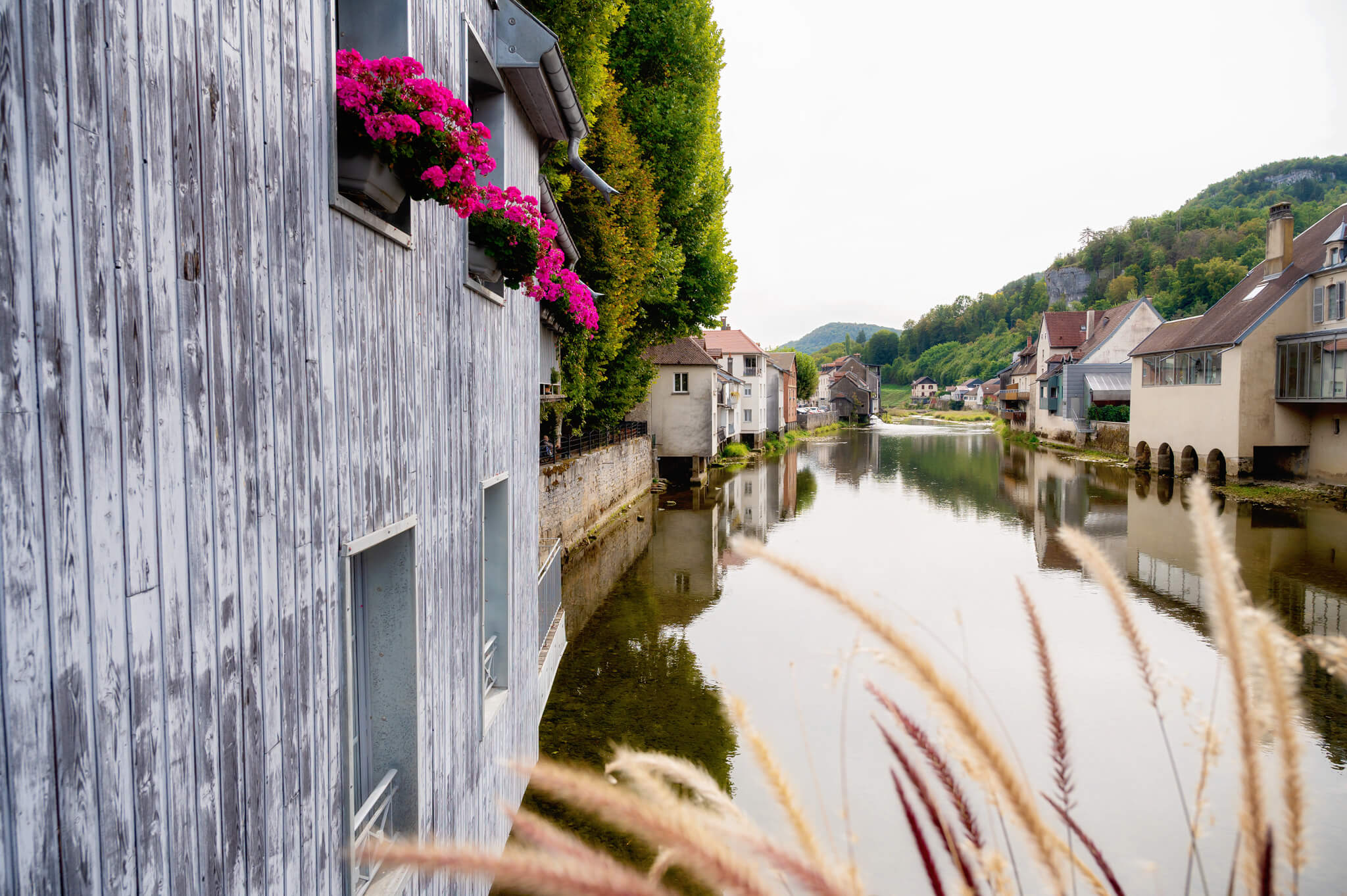 Ornans, France Grey wooden houses line a calm river, pink flower boxes in the windows and green hills in the background.