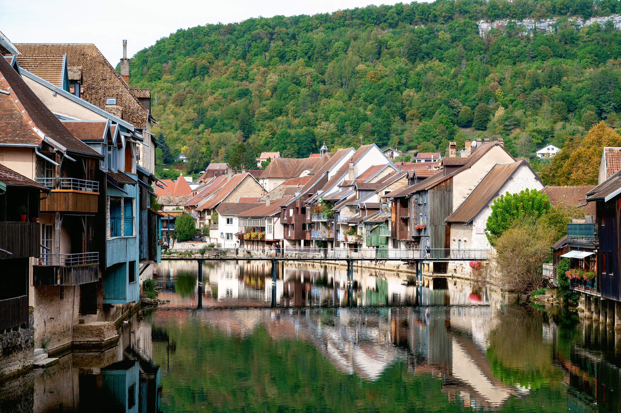 Ornans, France Riverside village with timber houses along a calm canal and a footbridge, framed by green forested hills in the background.