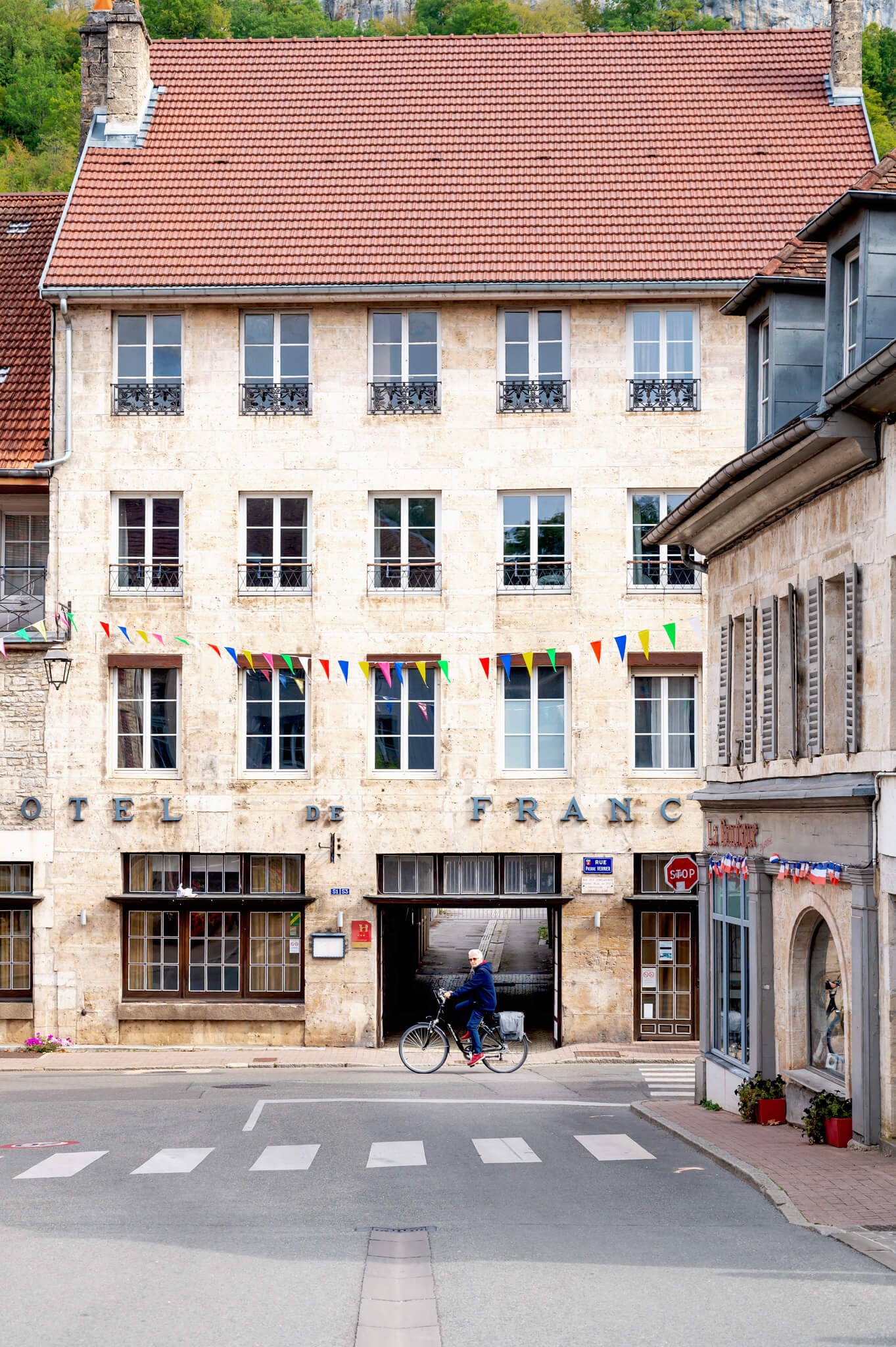 Ornans, France Stone hotel building with a red tiled roof, wrought-iron balconies, and colorful pennant banners spanning the street; a cyclist rides past the arched entrance.
