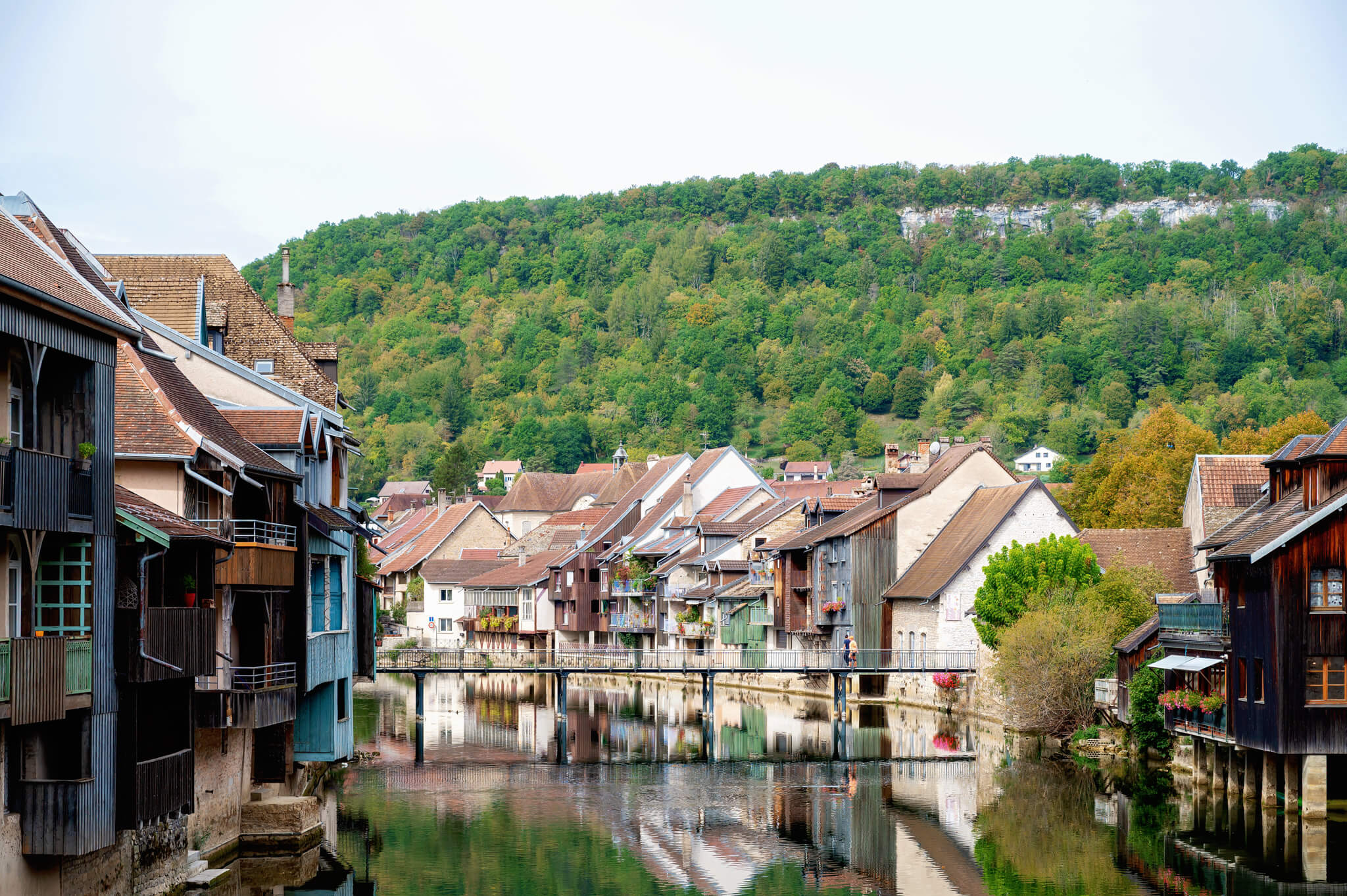 Ornans, France Row houses built over a calm river with reflections, and a forested hillside in the background.
