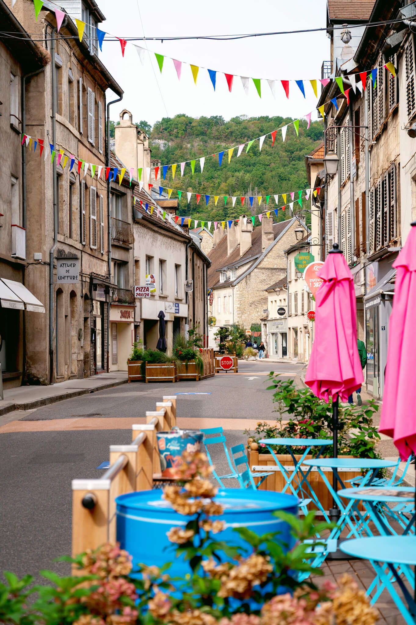 Ornans, France Colorful pennant banners strung above a European street with outdoor cafe tables and blue chairs beneath them.