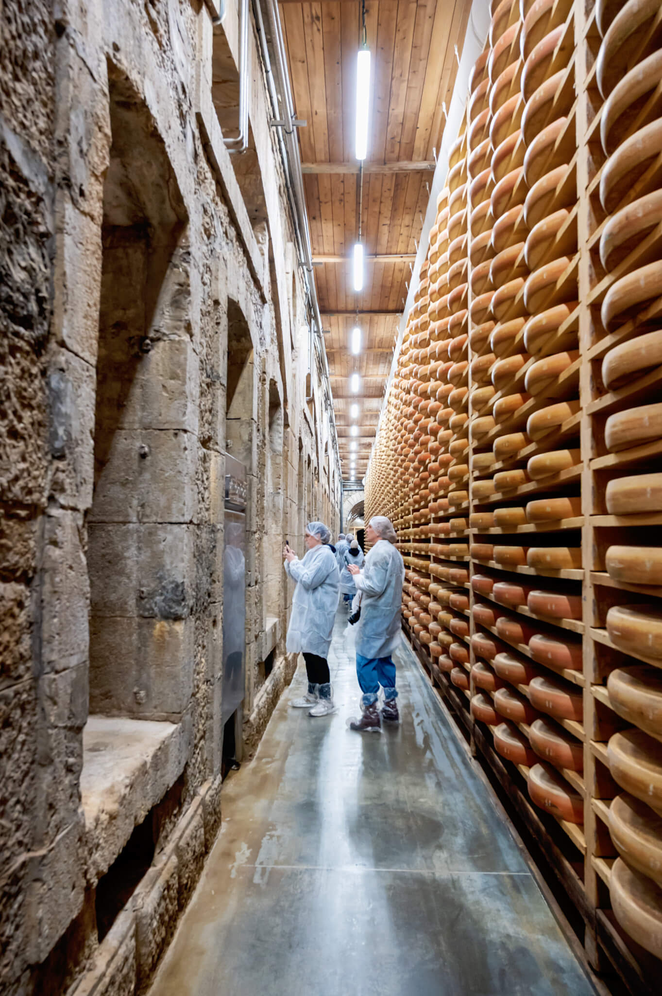 Visitors in protective coats walk between rough stone walls and tall racks of aging comte cheese wheels on the right in a narrow cellar.
