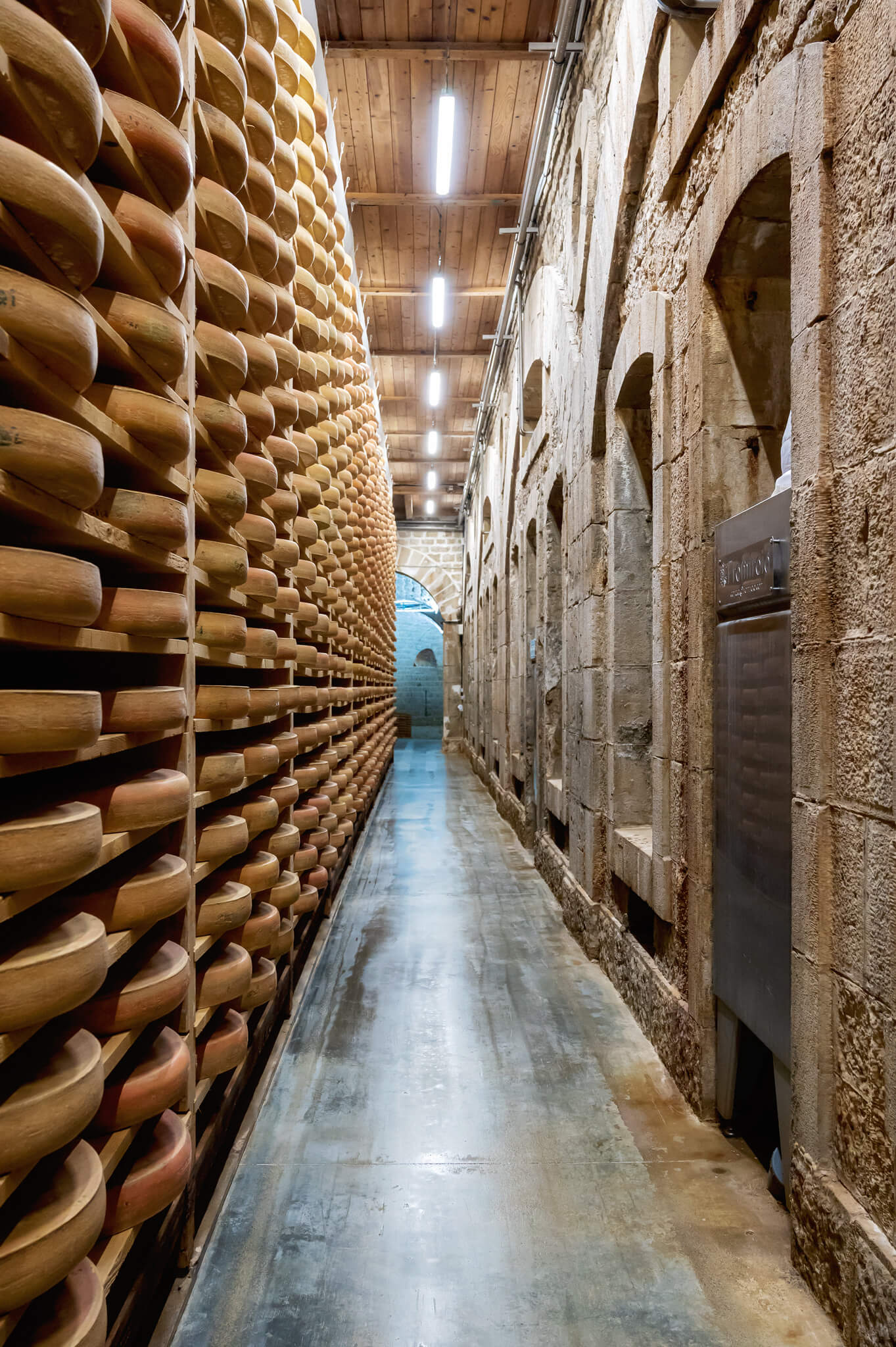 Narrow comte cellar corridor with tall wooden racks filled with round comte cheese wheels on the left and a stone wall with arches on the right.