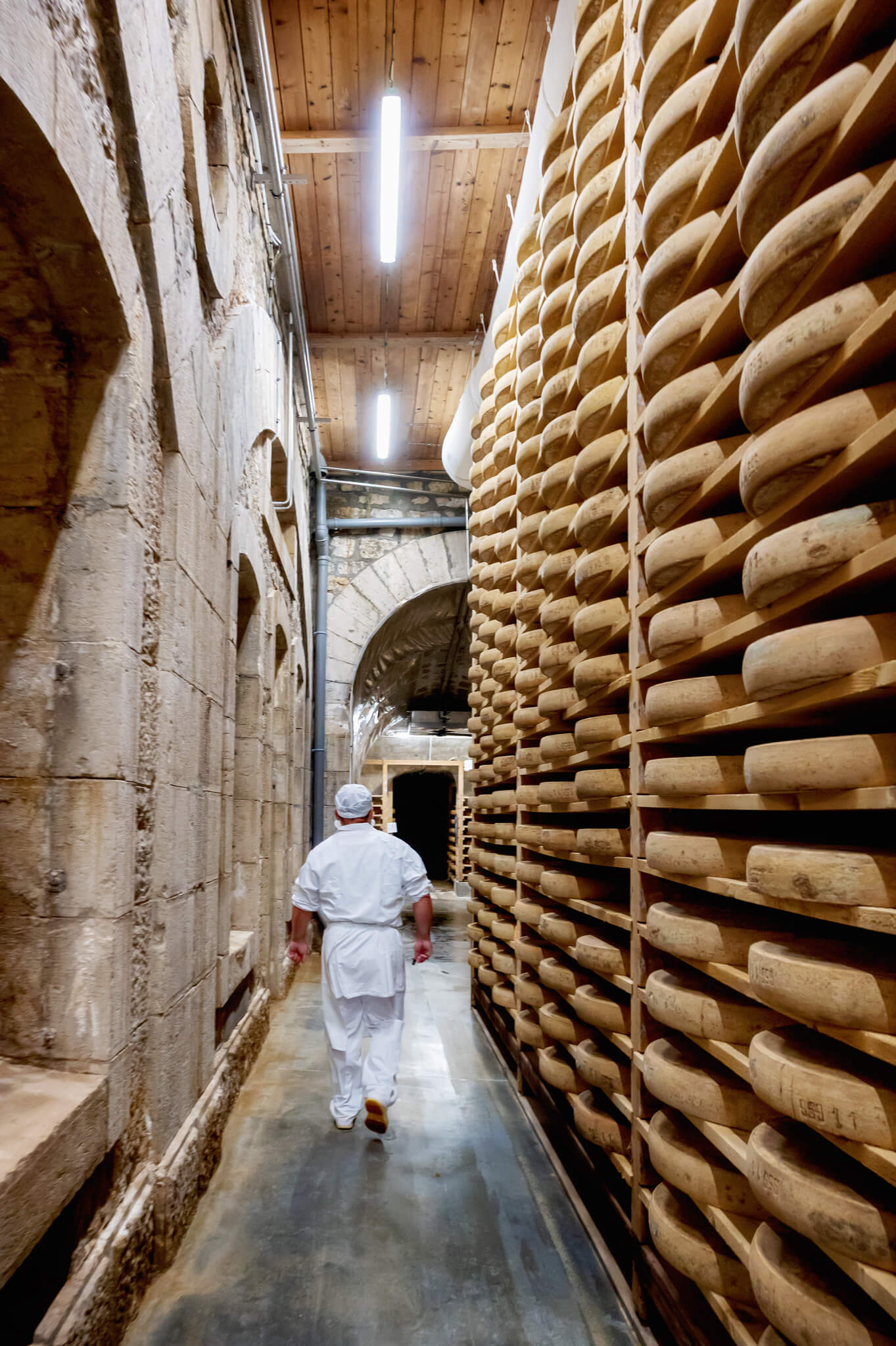 Worker in a white uniform walks down a narrow, stone-walled corridor flanked by towering shelves of aging comte cheese wheels on the right.
