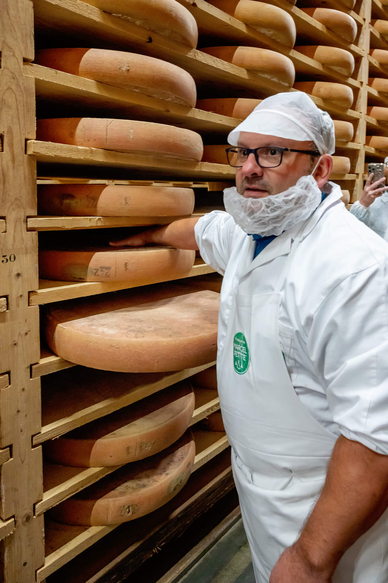 Worker in a white coat and hairnet stands beside aging racks of large comte cheese wheels, touching a wheel with gloved hand if needed.