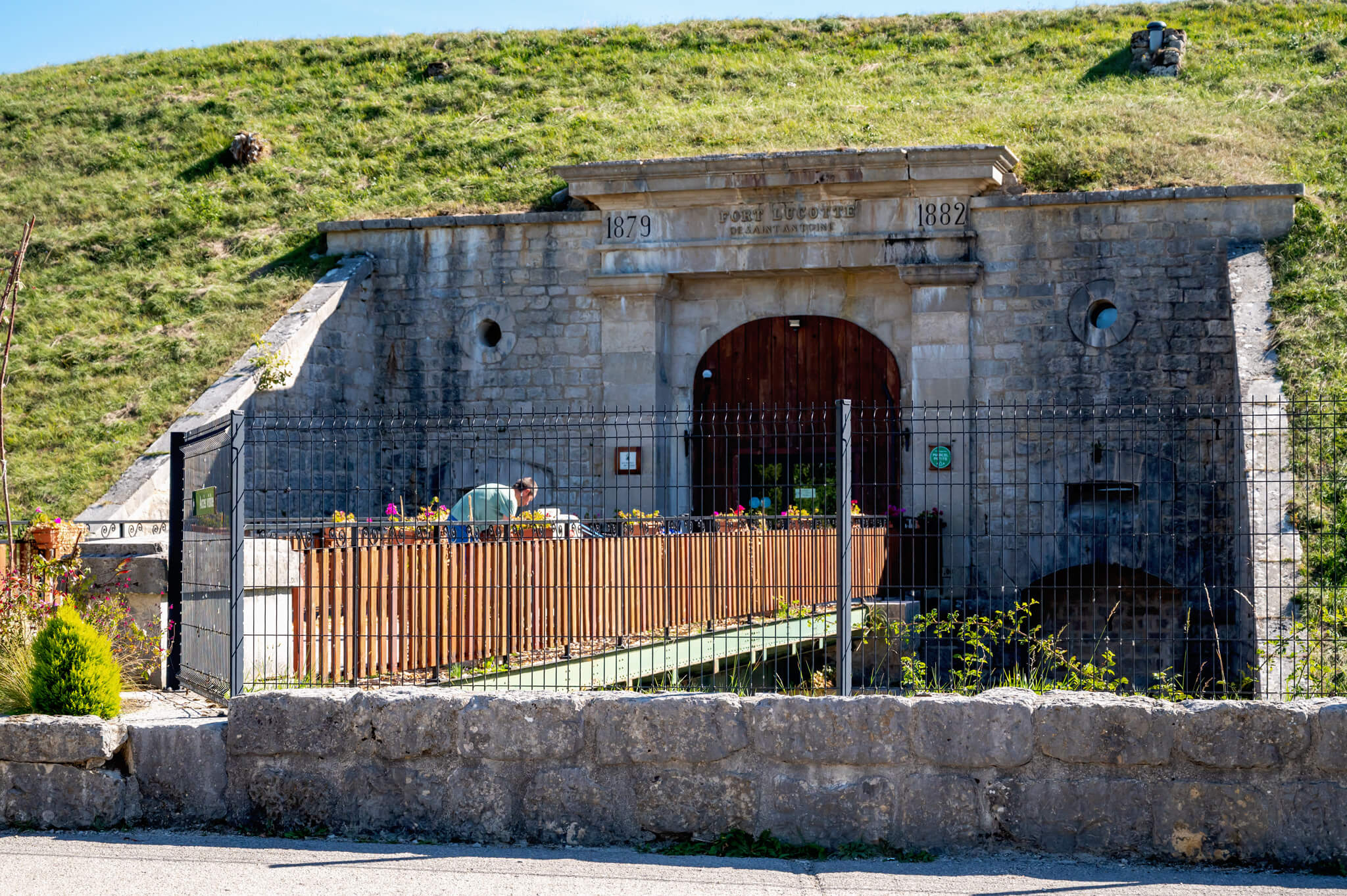Stone fort entrance labeled Fort Lucotte with the dates 1879 and 1882, built into a grassy hillside, behind a black metal fence.