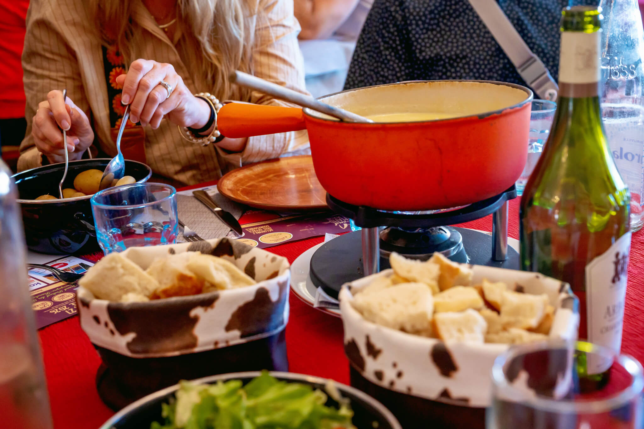 People dipping food at a table with a large orange comte cheese fondue pot and bread pieces on the side.