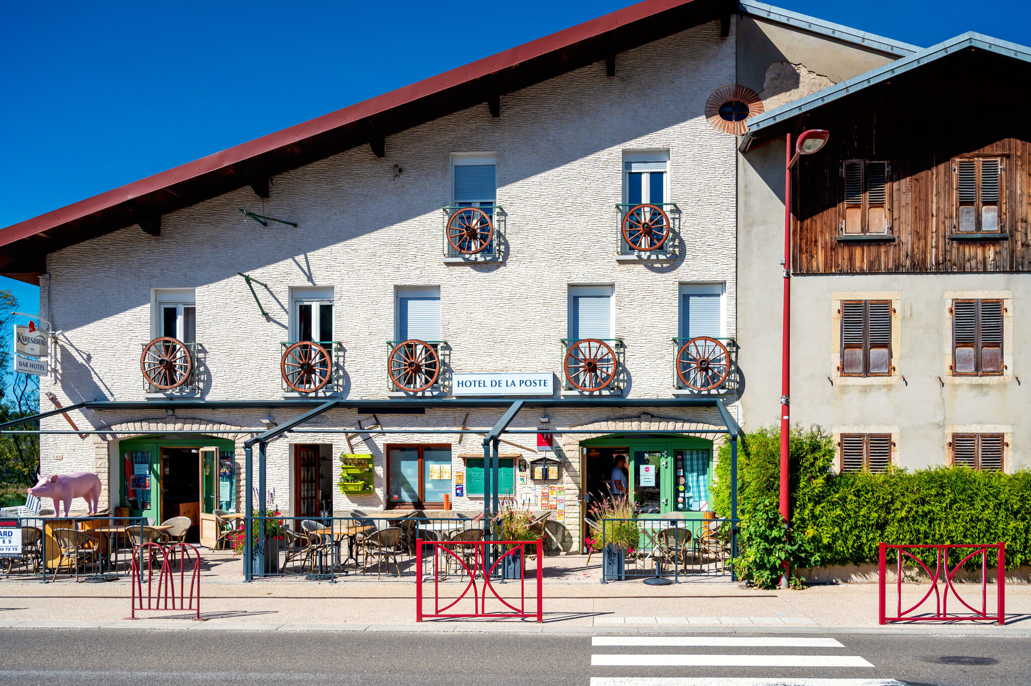 Hotel de la Poste building with white brick facade, red wagon-wheel balconies, and outdoor cafe seating under a clear blue sky.