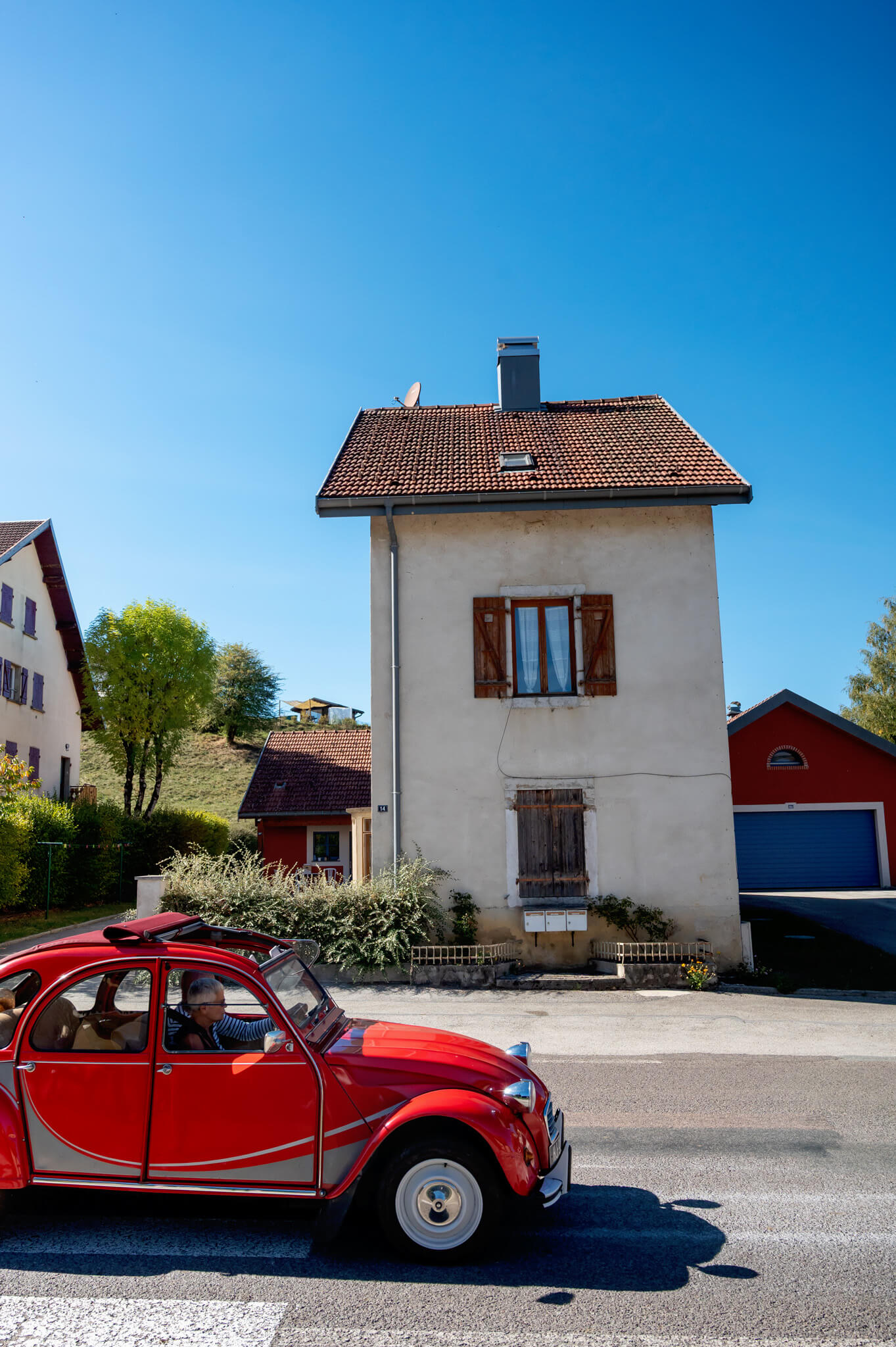 Red vintage convertible with two occupants driving along a sunny street in front of a beige house with wooden shutters and a tiled roof under a bright blue sky